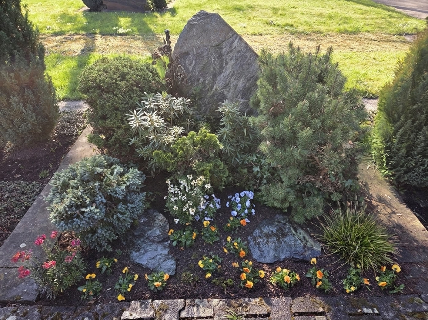 Dauergrabpflege mit Koniferen, Stauden und Hornveilchen an Natursteinfindling